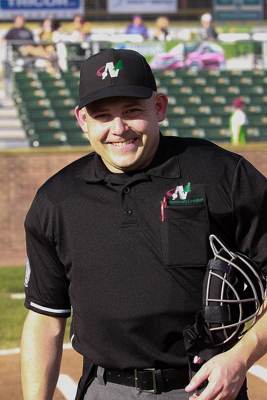 Kevin Macho in umpire uniform on a baseball field, smiling, mask under his arm